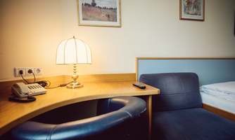 The combined work and seating area in the Small Room offers a functional desk with a lamp as well as a comfortable seating option. Classic furniture and warm colors create a cozy atmosphere – ideal for short stays or relaxed working in the unrenovated room of Hotel Hecher.