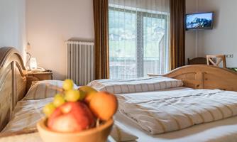 A cozy bedroom with two beds, a window, and a television. On the table, there is a fruit bowl with apples and grapes.