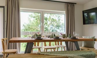 A modern dining area with a large wooden table and chairs. Daylight streams through the windows onto the elegant decor.