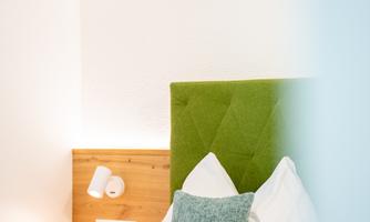 A bright bedroom with a cozy bed and a green headboard. On the bed lies a pillow as well as a modern wall lamp.