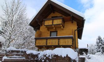 A pretty wooden house with a snow-covered roof. The surroundings are wintry with many snow-covered trees.