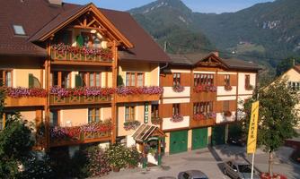 A picturesque hotel building with balconies full of flowers. In the background, green mountains rise beneath a clear sky.