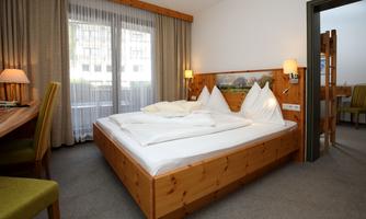 A cozy bedroom with a large double bed and two pillows. The room features large windows with curtains and wooden decoration.