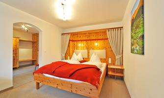 A cozy bedroom with a wooden bed and red pillows. The walls are painted in warm tones, and there are large windows with curtains.