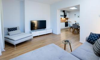 A modern living room with a large TV and a comfortable sofa. In the background, there is a dining area with a table and chairs.