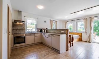 A modern kitchen with wooden flooring and light walls. Large windows let in plenty of light and provide a view of the greenery.