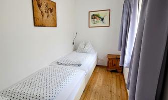 A simple bedroom with two beds and a wooden floor. Two pictures hang on the wall, and there are curtains at a window.