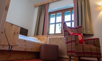 A cozy bedroom with wooden details and a large window. A comfortable armchair and a small stool complement the decor.
