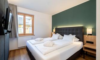 A modern bedroom with a double bed and two pillows. The room has large windows and a green wall.