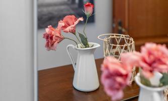 A white jug with pink flowers is placed on a wooden table. In the background, a decorative structure and a blurred image are visible.