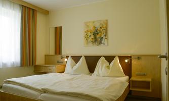 A cozy bedroom with a double bed and two pillows. A flower picture hangs on the wall, and there is a window with curtains.