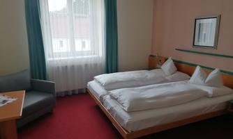 A simple hotel room with two single beds and a cozy armchair. The room has a red carpet and large windows that let in plenty of daylight.