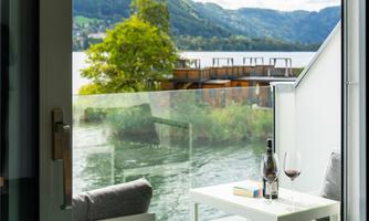 A beautiful view of a lake surrounded by mountains. On the terrace, there is a table and two chairs, with a bottle of wine beside them.