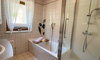 A modern bathroom with a bathtub and a separate shower. The windows are decorated with curtains, and a few plants provide a fresh atmosphere.