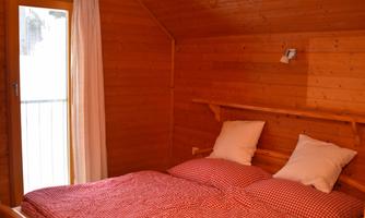 A cozy bedroom with wooden walls and a large double bed. The bedding is red and white checked, and there is a window with a curtain.