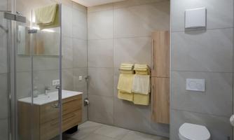 A modern bathroom with bright tiles and a glass shower enclosure. The room features a wash area with a wooden cabinet and yellow towels.