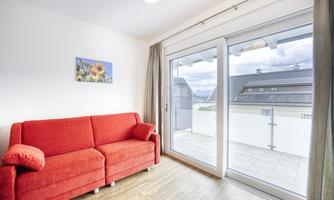 A bright room with a red sofa and a large window. The view looks out onto a balcony and a cloud landscape.
