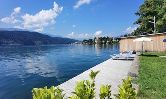 A calm lake with clear water and a blue sky. There are loungers and a sunshade at the shore, surrounded by trees.