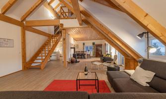 A bright, modern living room with wooden beams and a cozy sofa. A staircase leads to the upper area, where additional living spaces are located.