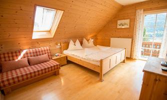 A cozy bedroom with wooden paneling, a double bed, and a sofa. Large windows provide plenty of daylight and a view outside.