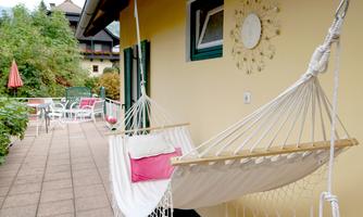 A cozy porch with a hammock and colorful cushions. In the background, garden furniture and a green environment can be seen.