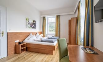 A cozy hotel room with a double bed, a desk, and a chair. The large windows provide plenty of natural light.