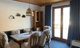 A cozy dining area with a large table and chairs adorned with fur covers. The windows let in plenty of daylight and offer a view outside.