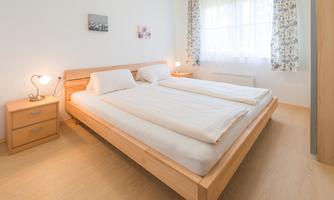 A bright bedroom with a large double bed and two nightstands. The windows are adorned with curtains, creating a pleasant light atmosphere.
