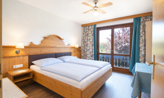 A cozy bedroom with a large double bed and wood paneling. The room has large windows overlooking nature and is cheerfully decorated.