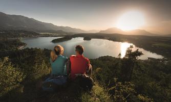 Two people are sitting on a hill and looking at a lake at sunset. The landscape features mountains in the background and green shores.