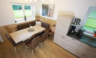A bright dining room with a large table and brown chairs. The modern kitchen is well-equipped and has large windows for plenty of daylight.