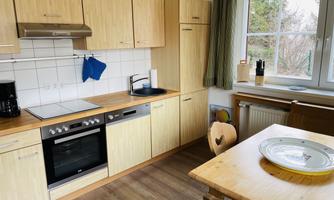 A modern kitchen with wooden cabinets and a large window. The dining table is simple and has a plate decoration.