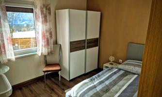 A simple bedroom with a bed, a wardrobe, and a window. The walls are light brown and the floor is made of wood.