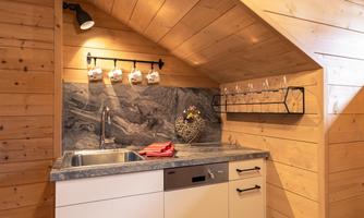 A cozy kitchen with wooden walls and a modern countertop. Cups hang on the wall and there are glasses on a shelf.