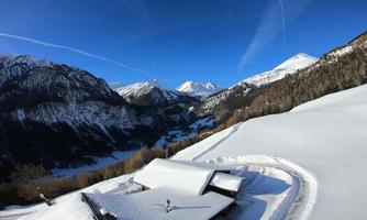 A snowy landscape with mountains and a clear blue sky. In the foreground, there is a house surrounded by snow-covered terrain.
