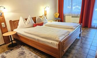 A cozy bedroom with a wooden double bed and red curtains. The walls are light, and there is a small table next to the bed.