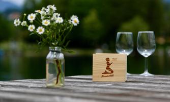 A rustic table with a small glass vase containing fresh flowers. Next to it are two wine glasses and a small wooden box with an engraved design.