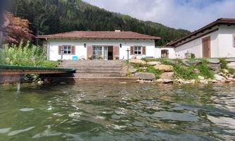 A charming house by the water with a spacious staircase and surrounding stones. In the background, green hills and trees can be seen.