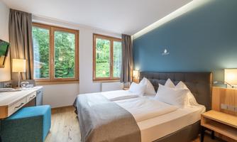 A bright hotel room with two beds and large windows. The walls are painted in a soft blue, and there are modern furnishings.