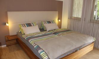 A cozy bedroom with a large double bed and colorful bedding. The room is bright and inviting, with large windows and wooden flooring.