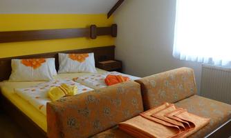 A cozy bedroom with a double bed and bright bedding. The walls are in yellow and brown, and there is a large window front for daylight.