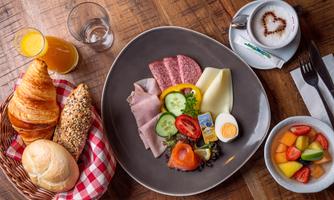 A lovingly arranged breakfast with sausage, cheese, vegetables, and an egg on a large plate. There are also rolls, croissants, and fresh fruit in a bowl, along with a drink.