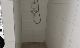 A modern shower with white tiles and a showerhead. The floor is light and a small towel lies on it.