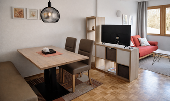 A modern living room with a dining table and chairs. In the background, there is a red sofa and a television on a shelf.