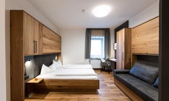 A modern hotel room with wooden paneling, two single beds, and a sofa bed. The large windows provide plenty of daylight.