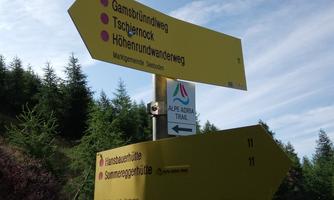A signpost with several hiking trails in the mountains. The surroundings consist of green forests and a clear sky.