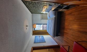 A modern bedroom with a double bed and wooden flooring. There are two red chairs and a large window door that lets in plenty of daylight.