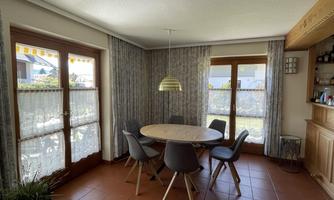 A bright dining room with a round table and six chairs. Large windows let in plenty of light and provide a view outside.