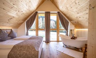 A cozy bedroom with wood walls and a large window. There is a modern bathtub and inviting bedding.
