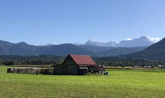 A rural scene with a barn and green meadows. In the background, majestic mountains and a clear blue sky can be seen.